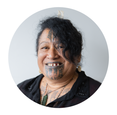 Tui is a Māori woman. She has a traditional moko kauae tattoo on her lips and chin. She also has a Māori design tattoo on her forehead. She's wearing a black shirt and pounamu (greenstone) pendant. The shirt is open a little to show that Tui's chest is al. 