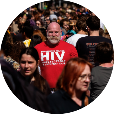 Mark Fisher is standing in a crowd, he is smiling and has a fabulous moustache. He is wearing a red t-shirt that says 'treatment makes HIV undetectable and uninfectious. . 