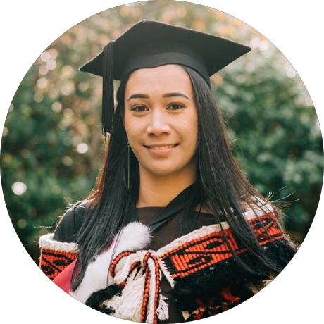 Sade is a young Māori wahine. She is facing the left, wearing a graduation cap and gown, with a korowai over the top. The korowai has koru detailing around the top, and is primarily black, red and white.  . 