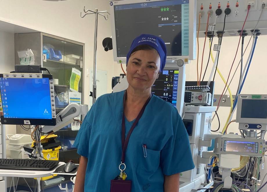 Dr Thomas stands in surgical scrubs in front of medical equipment. Her hat says "Dr Jane Anaesthetist". 