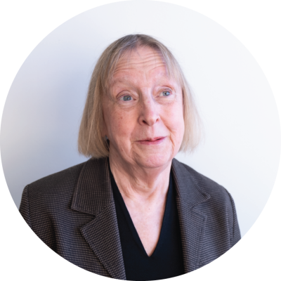 Mary Schnackenberg is an older Pākehā woman with a short blonde bob hair cut. Her smile looks like thinking of a great joke.. 