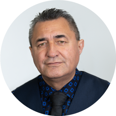 Trevor is a senior Māori man. He is looking at the camera with a warm smile. He is wearing a black suit blazer and a dark tie.. 